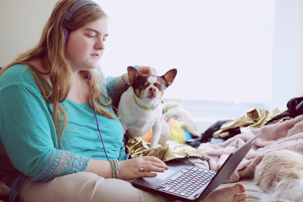 A young woman sitting on her bed working at a laptop. She is wearing headphones and has a small dog sitting next to her.